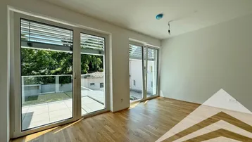 Helles Zimmer mit Parkettboden und Zugang zu einer großen gefliesten Terrasse mit Blick ins Grüne.
