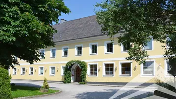Traditionelles gelbes Gebäude mit vielen Fenstern und einem begrünten Eingangsbereich unter blauem Himmel.