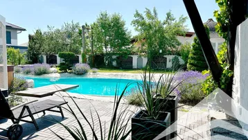 Großer Außenpoolbereich mit gepflasterter Terrasse, Sonnenliegen und üppiger Bepflanzung unter blauem Himmel.