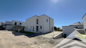 Moderne weiße Doppelhaushälfte mit Flachdach und Zufahrt unter strahlend blauem Himmel.