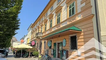 Historisches Gebäude mit Geschäften im Erdgeschoss und traditionellen Fensterläden an einer belebten Straße.