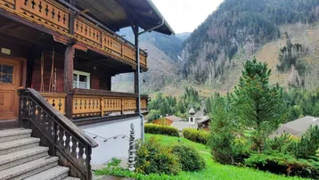 Traditionelles Holzhaus mit geschnitztem Balkon und Steintreppe, umgeben von alpiner Berglandschaft.