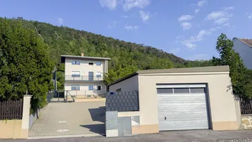Hausfassade mit Garage und Zufahrt, umgeben von grünen Hügeln unter blauem Himmel.
