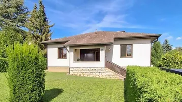 Einfamilienhaus mit weißer Fassade, umgeben von gepflegtem Rasen und grünen Hecken unter blauem Himmel.