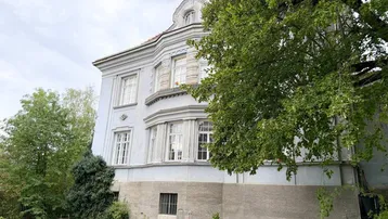 Historisches Gebäude mit klassischer Architektur und großen Fenstern, umgeben von Grün.