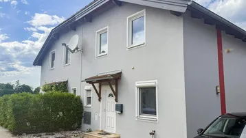 Graue Hausfassade mit weißen Fenstern, Haustür und Satellitenschüssel unter bewölktem Himmel.