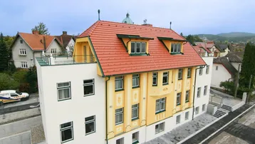 Gebäudeansicht von oben mit rotem Ziegeldach und gelber Fassade, umgeben von weiteren Häusern.