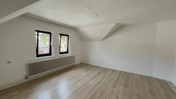 Helles Zimmer im Dachgeschoss mit Laminatboden und Heizkörper unter den Fenstern.