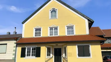 Gelbes Wohnhaus mit braunem Ziegeldach, grünen Fensterläden und weiß gerahmten Fenstern unter blauem Himmel.