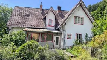 Altes, charmantes Haus mit weißer Fassade, roten Fensterrahmen und verwildertem Garten.