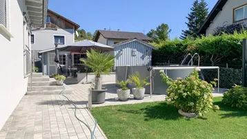 Gepflegter Gartenbereich mit Rasenfläche, gepflasterter Terrasse und einem Aufstellpool unter blauem Himmel.