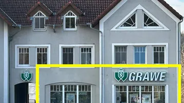 Graue Fassade eines Gebäudes mit mehreren Fenstern und einem Geschäft im Erdgeschoss, das Dach ist mit roten Ziegeln gedeckt.