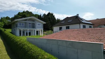 Haus mit Satteldach und Photovoltaikanlage, umgeben von Hecken und Grünflächen.