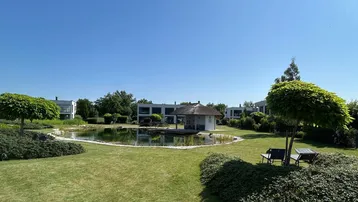 Großer, gepflegter Gartenbereich mit Teich, Sitzgelegenheiten und modernen Gebäuden im Hintergrund.