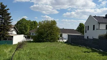 Weitläufiges Baugrundstück mit grüner Wiese und umliegenden Wohnhäusern unter blauem Himmel.