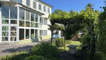 Grüner Gartenbereich mit Pergola, Sitzgelegenheit und Blick auf das Haus unter blauem Himmel.