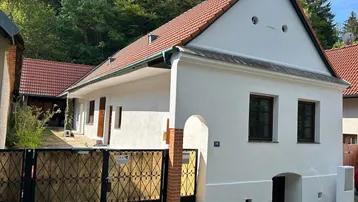 Weißes Haus mit rotem Ziegeldach und schmiedeeisernem Tor, umgeben von Bäumen.