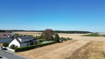 Ländliche Umgebung mit weiten Feldern, einigen Wohnhäusern und einer Hecke unter klarem Himmel.
