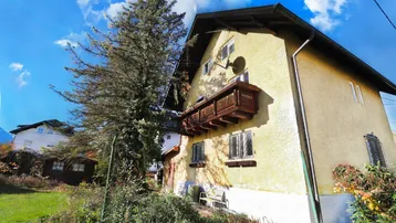 Traditionelles Haus mit gelber Putzfassade, Holzbalkon und umgebender Natur unter blauem Himmel.