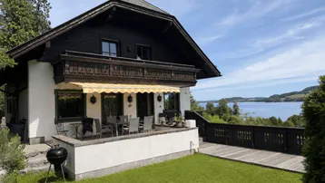 Traditionelles Haus mit großer Terrasse, Markise und herrlichem Seeblick unter blauem Himmel.