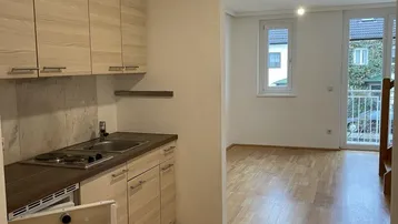 Helle offene Wohnfläche mit integrierter Küchenzeile, Parkettboden und Zugang zu einem Balkon.