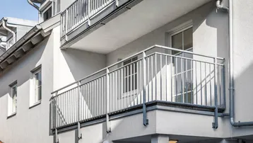 Balkon mit Metallgeländer an der Fassade eines Mehrfamilienhauses in hellem Grau.