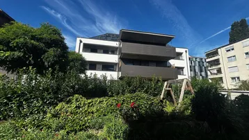 Außenansicht eines modernen Wohngebäudes mit Balkonen, umgeben von üppigem Grün und blauem Himmel.