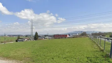 Grüne Wiese mit Blick auf eine Straße, Häuser und Berge im Hintergrund unter blauem Himmel.