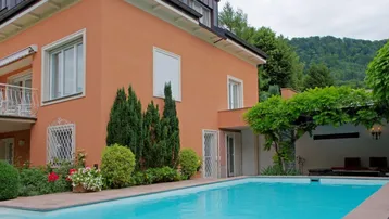 Hausfassade in Terrakottafarbe mit Swimmingpool und grüner Bepflanzung im Vordergrund, umgeben von Bergen.