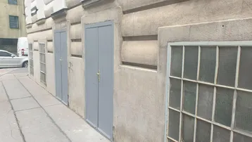 Alte Gebäudefassade auf Straßenniveau mit zwei grauen Türen und vergitterten Kellerfenstern.