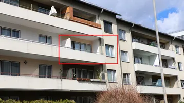 Blick auf ein Mehrfamilienhaus mit einem rot umrandeten Balkon unter bewölktem Himmel.
