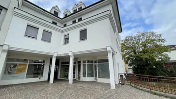 Modernes Geschäftsgebäude mit weißer Fassade, großen Fenstern und gepflastertem Vorplatz unter bewölktem Himmel.
