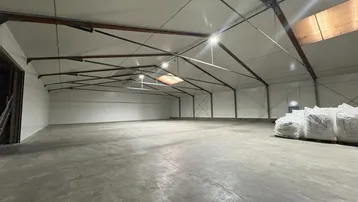 Geräumige, leere Lagerhalle mit Betonboden, hohen Decken und sichtbaren Dachbalken unter heller Beleuchtung.