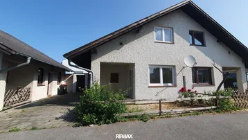 Einfamilienhaus mit heller Fassade, Satteldach und angrenzendem Nebengebäude unter blauem Himmel.