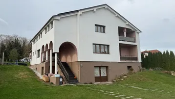 Großes Wohnhaus mit weißer Fassade, braunem Sockel und mehreren Balkonen auf einem grünen Hanggrundstück.
