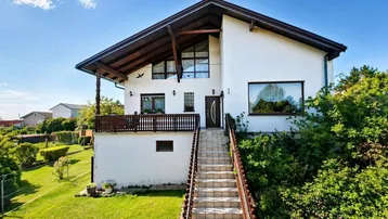 Weißes Einfamilienhaus mit Holzbalkon und langer Steintreppe zum Eingang.