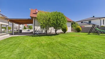 Großer Garten mit Rasenfläche, Bäumen, Spielplatz und überdachtem Stellplatz neben dem Haus.