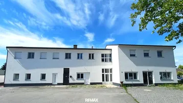 Weißes, modernes Gebäude mit vielen Fenstern und einem Parkplatz davor unter blauem Himmel.