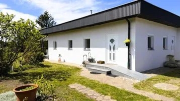Weißes Einfamilienhaus mit dunklem Dachüberstand, gepflegtem Vorgarten und barrierefreiem Zugang.