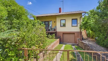 Gelbes Einfamilienhaus mit integrierter Garage, Balkon und üppiger Vegetation.