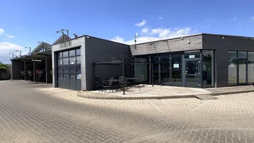 Modernes Geschäftsgebäude mit gepflastertem Vorplatz, Außenterrasse und angrenzenden Servicebereichen unter blauem Himmel.