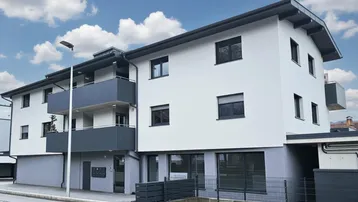 Modernes Mehrfamilienhaus mit weißer Fassade, grauen Akzenten und Balkonen unter blauem Himmel.