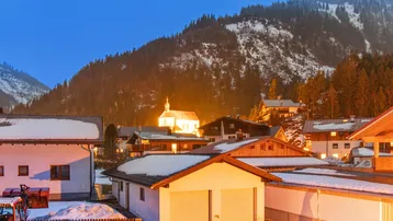 Winterliche Abendansicht eines Dorfes mit beleuchteten Häusern und schneebedeckten Bergen im Hintergrund.