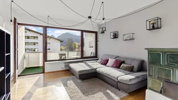 Helles Wohnzimmer mit gemütlichem Sofa, Kachelofen und Zugang zum Balkon mit Bergblick.