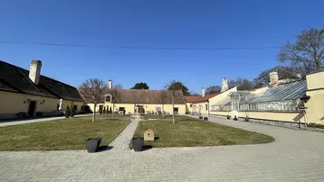 Weitläufige Außenansicht eines historischen Anwesens mit gepflastertem Hof, Rasenfläche und Gewächshaus unter blauem Himmel.