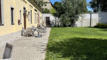 Gepflasterte Terrasse mit Gartenmöbeln und Rasenfläche neben einem historischen Gebäude unter blauem Himmel.