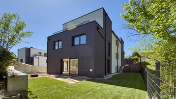Modernes Reihenhaus mit dunkler Fassade und direktem Zugang zum grünen Gartenbereich unter blauem Himmel.