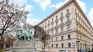 Historisches Gebäude mit klassischer Architektur und einer Statue auf dem Vorplatz unter blauem Himmel.