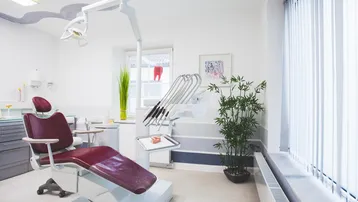 Heller Behandlungsraum mit moderner Zahnarztliege, großen Fenstern und Jalousien für Privatsphäre.