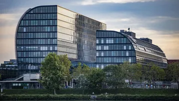Modernes Bürogebäude mit geschwungener Glasfassade am Wasser, umgeben von Bäumen und Grünflächen.